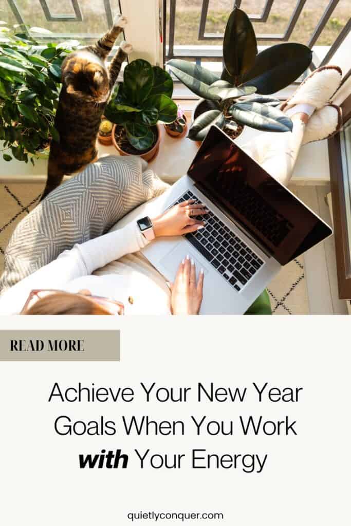 Woman sat by window, cat near her, working on macbook, sun shining in. Text reads "Achieve your New Year Goals When You Work With Your Energy"
