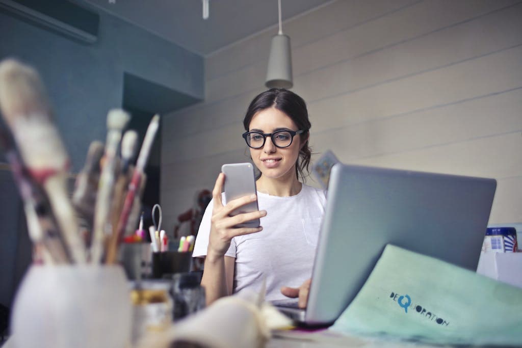 Digital Products for Growth: A woman using her phone at a desk, surrounded by art supplies and a laptop, in a creative workspace.