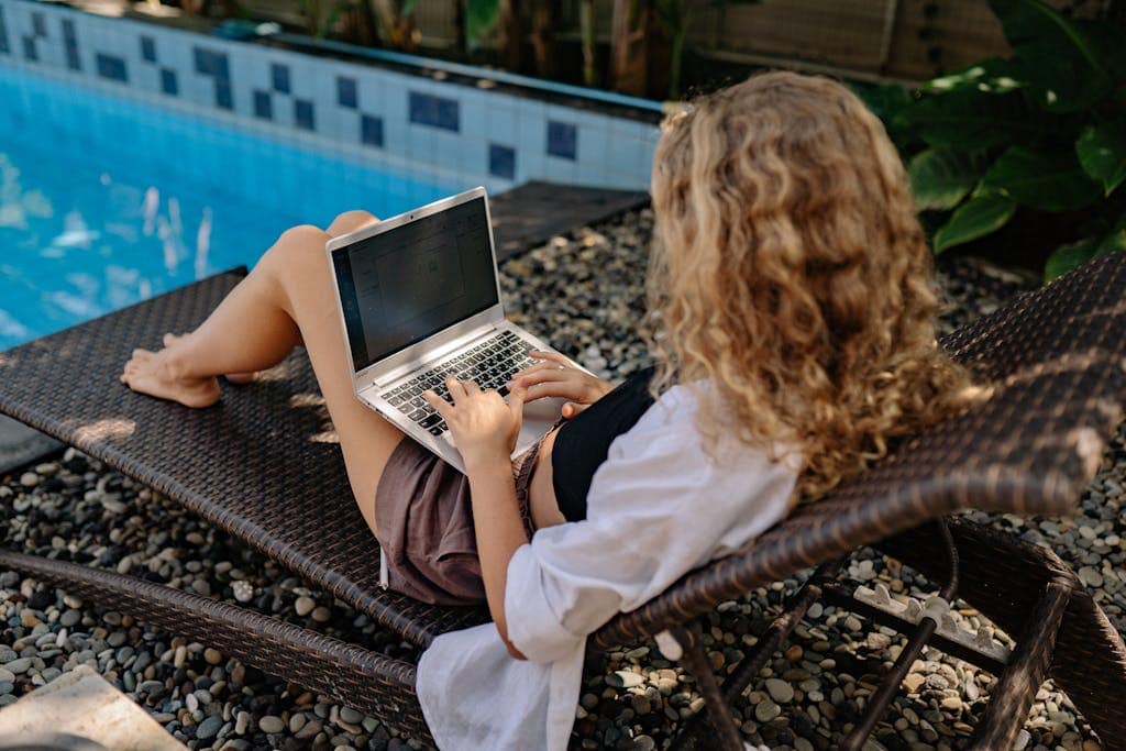A woman with curly hair works on a laptop while lounging by the pool in a relaxed outdoor setting.