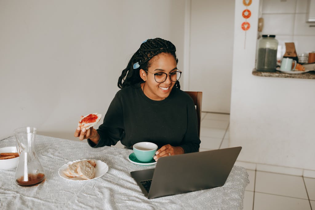 Young woman having breakfast while working on her laptop at home, smiling and relaxed.
