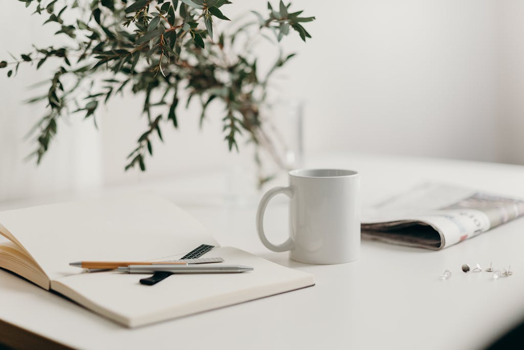 A serene minimalist workspace featuring a white coffee mug, open notebook, and green plant vase on a desk.